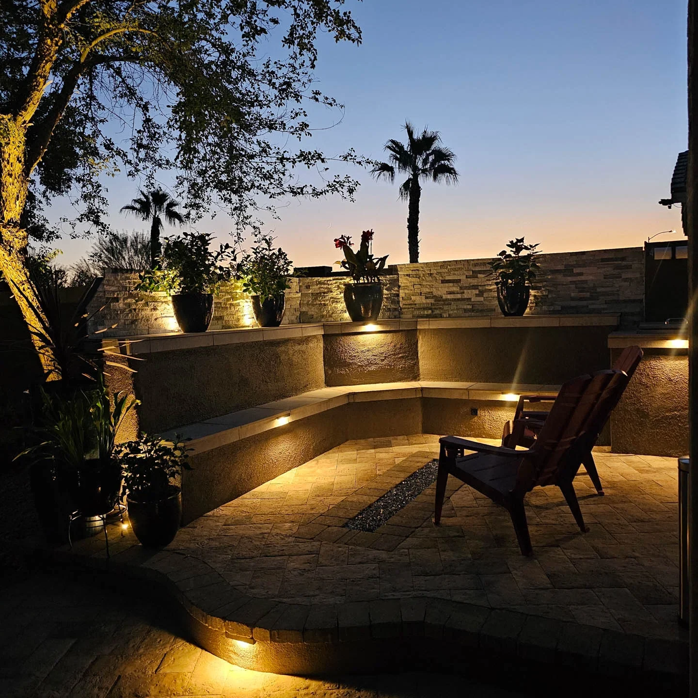 Illuminated backyard patio with chair, palms, and potted plants at dusk