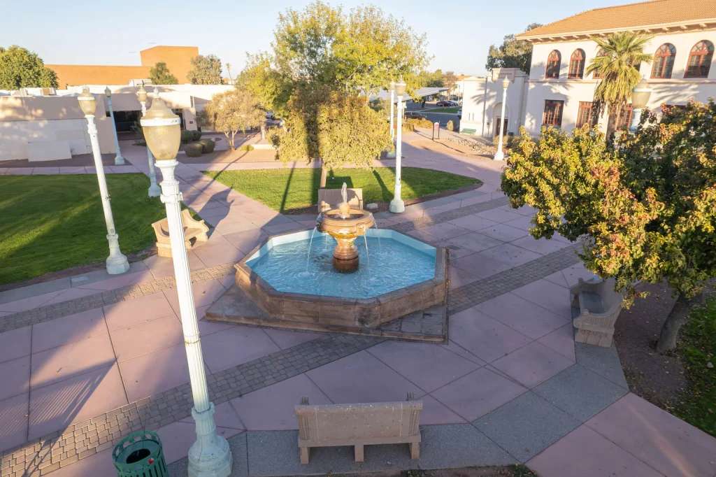 Sunlit courtyard fountain surrounded by trees, benches, and buildings